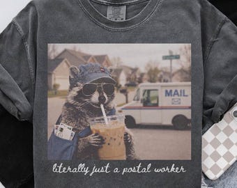 May include: A charcoal gray t-shirt featuring a graphic of a raccoon wearing a blue baseball cap and sunglasses, holding a plastic cup with a straw. The raccoon carries a blue mail bag. Below the image, the text reads, literally just a postal worker.