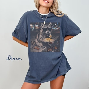 May include: A denim-colored short-sleeve t-shirt featuring a graphic print of a raccoon wearing black sunglasses and holding a plate of deviled eggs. The raccoon is positioned in an urban setting at night with blurred city lights in the background.