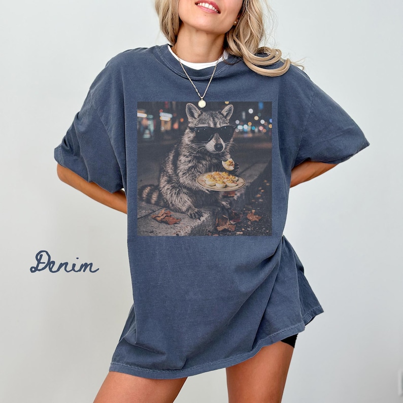May include: A denim-colored short-sleeve t-shirt featuring a graphic print of a raccoon wearing black sunglasses and holding a plate of deviled eggs. The raccoon is positioned in an urban setting at night with blurred city lights in the background.