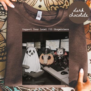 May include: A dark chocolate crewneck sweatshirt featuring a graphic print with the text Support Your Local 911 Dispatchers. The image shows three animals wearing headsets at a desk, dressed as a ghost, a pumpkin, and a vampire.
