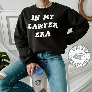 May include: Black sweatshirt with white text that reads "In My Lawyer Era". The sweatshirt is a unisex style and has a wave graphic on the bottom left corner.