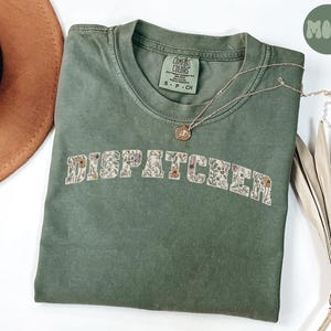May include: A green t-shirt with the word "DISPATCHER" in floral lettering. The shirt is made from a soft, comfortable fabric and has a relaxed fit.