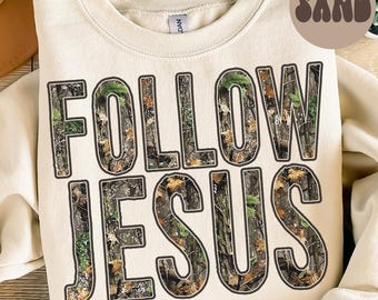 Puede incluir: Una sudadera de color arena con las palabras "FOLLOW JESUS" en letras grandes y en negrita. Las letras están rellenas con un patrón de camuflaje verde, marrón y beige. La palabra "SAND" está en un círculo marrón en la esquina superior derecha.