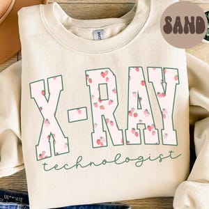 Può includere: Una felpa color sabbia con la scritta "X-RAY technologist". Le lettere sono bordate di verde e riempite con un motivo a fragole rosa. La parola "SAND" è stampata in un cerchio marrone in alto a destra.