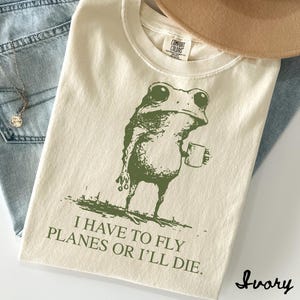 May include: Ivory-colored t-shirt with a green frog graphic holding a mug. The text "I HAVE TO FLY PLANES OR I'LL DIE" is printed below the frog. The shirt is folded on top of blue jeans.