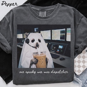 Peut inclure: Un t-shirt à col rond gris anthracite avec un motif représentant un raton laveur déguisé en fantôme avec un drap blanc et un casque, tenant un café glacé. L'image montre un centre de répartition. Le texte indique : one spooky wee woo dispatcher.