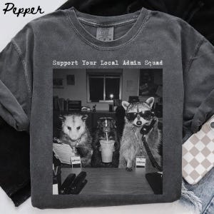May include: A charcoal gray t-shirt featuring a black and white graphic of an opossum, a skunk, and a raccoon sitting at an office desk. The raccoon wears sunglasses and holds a telephone receiver. The text reads Support Your Local Admin Squad.
