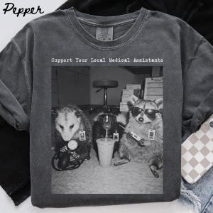 May include: A charcoal grey t-shirt featuring a black and white graphic of an opossum, a skunk, and a raccoon wearing medical ID badges. The raccoon wears sunglasses and holds a thermometer. The text reads Support Your Local Medical Assistants.