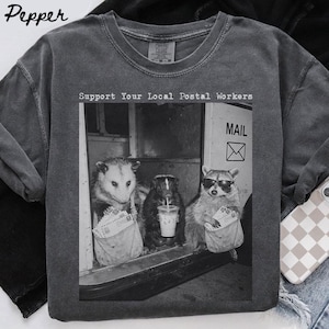 May include: A charcoal grey crew neck t-shirt featuring a black and white graphic of an opossum, a skunk, and a raccoon wearing sunglasses, all holding mail bags. The text above the graphic reads, Support Your Local Postal Workers.