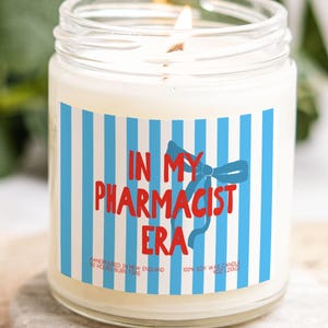 May include: A lit soy wax candle in a clear glass jar. The label features blue and white vertical stripes, with the text "IN MY PHARMACIST ERA" in red and a blue bow. The candle is 113g and hand-poured in New England.