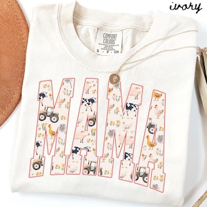A white t-shirt with a large text design that says "MAMA" printed on it, featuring various farm and animal-themed illustrations.