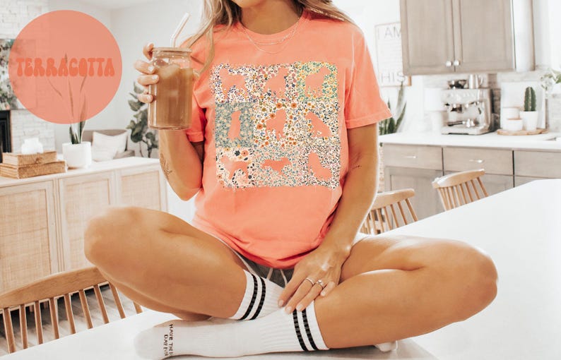 Op de afbeelding: Terracotta-kleurig T-shirt met een bloemen- en dierenprint. Het shirt heeft een zachte, koraalkleur. Het woord "TERRACOTTA" is gedrukt op een grote, ronde, koraalkleurige achtergrond.