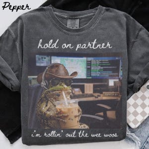 May include: A charcoal grey crew neck t-shirt featuring a graphic of a frog wearing a brown cowboy hat, sunglasses, and a headset while holding an iced coffee. The text reads hold on partner above the image and i'm rollin' out the wee woos below.