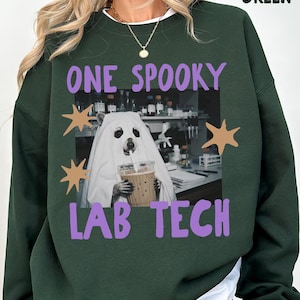 Könnte beinhalten: Ein waldgrüner Rundhals-Pullover mit einem Aufdruck eines Bären im weißen Geisterkostüm, der einen Eiskaffee in einem Labor hält. Der Text lautet ONE SPOOKY LAB TECH in Lila, ergänzt durch beigefarbene Sternformen an den Seiten.