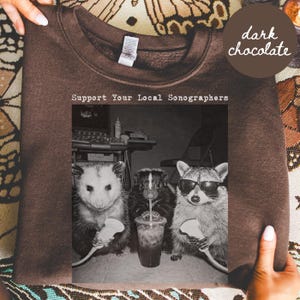 May include: A dark chocolate brown crewneck sweatshirt featuring a black and white graphic print. The text reads Support Your Local Sonographers above an image of an opossum, a skunk, and a raccoon wearing sunglasses, each holding a medical probe.