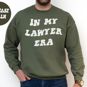 May include: A military green sweatshirt with the text "In My Lawyer Era" in white, retro-style lettering.