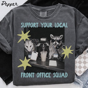Könnte beinhalten: Ein anthrazitfarbenes T-Shirt mit einer Grafik von drei Tieren an einem Büroschreibtisch. Der Text lautet SUPPORT YOUR LOCAL FRONT OFFICE SQUAD. Die Grafik zeigt einen Waschbären mit Sonnenbrille, ein Opossum und ein Stinktier mit gelben Sternen.