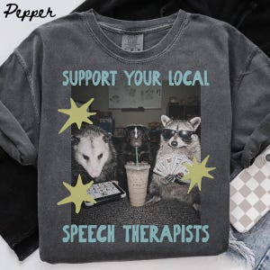 Op de afbeelding: Een antracietgrijs t-shirt met ronde hals en een grafische print van een opossum, een stinkdier en een wasbeer. De wasbeer draagt een zonnebril en houdt een waaier van kaarten vast. De tekst luidt SUPPORT YOUR LOCAL SPEECH THERAPISTS met gele sterren.