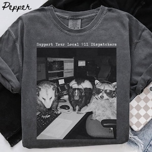 Puede incluir: Una camiseta de cuello redondo gris carbón con un estampado gráfico en blanco y negro de una zarigüeya, una mofeta y un mapache con auriculares en una estación de trabajo. El texto sobre la imagen dice Support Your Local 911 Dispatchers.