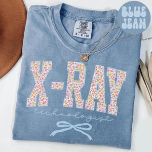 Op de afbeelding: Blauw spijkerbroekkleurig T-shirt met de woorden "X-RAY" in grote letters met bloemenprint. Onder de woorden "X-RAY" staat het woord "technologist" in script. Onderaan het T-shirt zit een lichtblauwe strik.