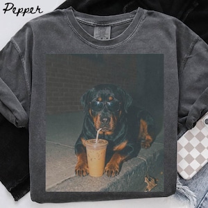 May include: A charcoal gray crew neck t-shirt featuring a graphic print of a Rottweiler dog wearing black sunglasses and drinking from a clear plastic cup with a straw. The shirt has a faded, vintage-style wash and the word Pepper is printed.