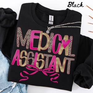 Puede incluir: Sudadera negra con la frase "MEDICAL ASSISTANT" en rosa y estampado de leopardo. Un corazón y un lazo rosas acentúan el diseño. También se ve un collar plateado con un colgante de flor.