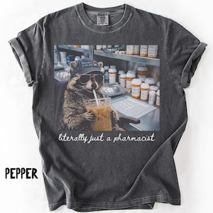 May include: Pepper-colored t-shirt featuring a raccoon wearing sunglasses and a pharmacy hat, drinking an iced coffee. The image includes the text "literally just a pharmacist". The shirt has rolled-up sleeves.