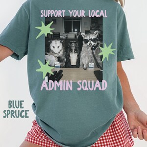 May include: A blue spruce t-shirt featuring a graphic of an opossum, a skunk, and a raccoon in an office setting. The text reads SUPPORT YOUR LOCAL ADMIN SQUAD in pink letters. The design includes light green star shapes around the central image.