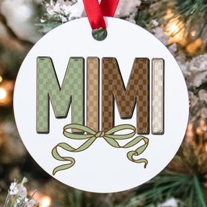 Puede incluir: Adorno navideño redondo blanco con la palabra "MIMI" en letras grandes a cuadros. Las letras son en tonos verdes, marrones y beige. Una cinta verde en la parte inferior y una cinta roja en la parte superior para colgar. Decoración festiva.