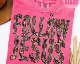 Puede incluir: Una camiseta rosa Comfort Colors con las palabras "FOLLOW JESUS" en letras grandes con estampado de camuflaje. Se ve un collar dorado con un colgante. La camiseta está doblada y un sombrero marrón es parcialmente visible en el fondo.