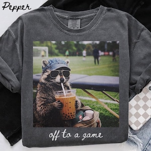 May include: Dark gray sweatshirt featuring a raccoon wearing sunglasses and a denim cap, drinking a beverage with a straw. The image has the text "off to a game" at the bottom. A small bag with medical supplies is visible.