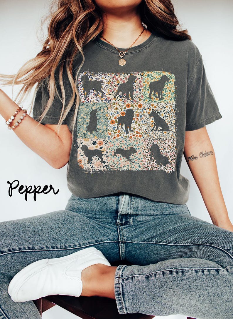 Op de afbeelding: Donkergrijs T-shirt met een bloemenhond silhouet ontwerp. Het shirt heeft een ronde hals en korte mouwen. Het woord "Pepper" staat cursief links onder in de afbeelding. Het model draagt een blauwe jeans en witte sneakers.