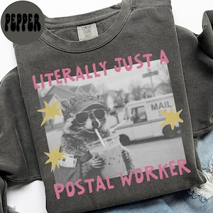 Könnte beinhalten: Ein anthrazitfarbenes Sweatshirt mit Rundhalsausschnitt und einem Aufdruck eines Waschbären mit Sonnenbrille und Kappe, der ein Getränk mit Strohhalm hält. Der Text LITERALLY JUST A POSTAL WORKER ist rosa, ergänzt durch gelbe Sterne und ein Postauto.