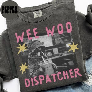 May include: A charcoal grey crewneck sweatshirt featuring a graphic print of a raccoon wearing sunglasses and a headset while holding a drink. The text WEE WOO DISPATCHER is printed in pink above and below the image, with yellow star accents.