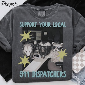 Puede incluir: Una camiseta gris carbón con un gráfico de una zarigüeya, una mofeta y un mapache con auriculares en estaciones de trabajo. El texto dice SUPPORT YOUR LOCAL 911 DISPATCHERS en azul claro, acentuado por dos formas de estrella de color amarillo.