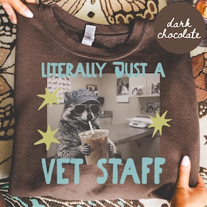 May include: A dark chocolate brown crewneck sweatshirt featuring a graphic of a raccoon wearing a baseball cap and sunglasses while holding an iced coffee. The text reads LITERALLY JUST A VET STAFF with light green star accents surrounding the image.