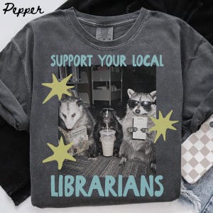 Puede incluir: Una camiseta gris carbón con un gráfico de una zarigüeya, una mofeta y un mapache en una biblioteca. El texto "SUPPORT YOUR LOCAL LIBRARIANS" aparece en azul claro. Los animales sostienen un libro, una bebida y un bloc de notas con estrellas amarillas.