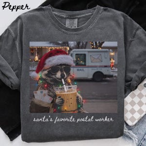 May include: A charcoal grey t-shirt featuring a graphic of a raccoon wearing a Santa hat and sunglasses, holding an iced coffee with a straw. The raccoon wears a mail bag with letters and is wrapped in Christmas lights. Text reads: santa's favorite postal worker.