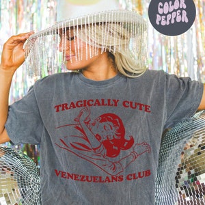 May include: A gray t-shirt with the text "Tragically Cute" and "Venezuelans Club" in red. A cartoon image of a woman with red hair is in the center of the shirt.