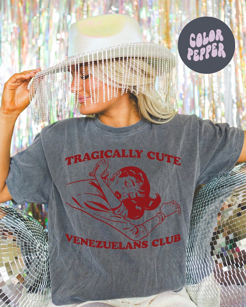 May include: A gray t-shirt with the text "Tragically Cute" and "Venezuelans Club" in red. A cartoon image of a woman with red hair is in the center of the shirt.
