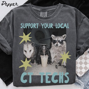 May include: A charcoal gray t-shirt featuring a graphic print of an opossum, a skunk, and a raccoon wearing sunglasses. The raccoon holds a plastic cup with a straw. The text reads SUPPORT YOUR LOCAL CT TECHS in light blue with yellow star accents.