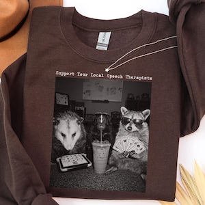 May include: A dark chocolate brown crewneck sweatshirt featuring a black and white graphic print of an opossum, a skunk, and a raccoon playing cards and drinking iced coffee. The text above the graphic reads, Support Your Local Speech Therapists.