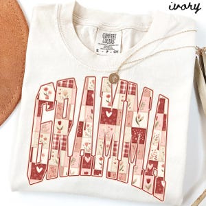 May include: Ivory-colored t-shirt with the word "GRAMMA" in a patchwork design of red, pink, and white squares. The squares feature floral and heart patterns. A gold necklace with a pendant rests on the shirt.