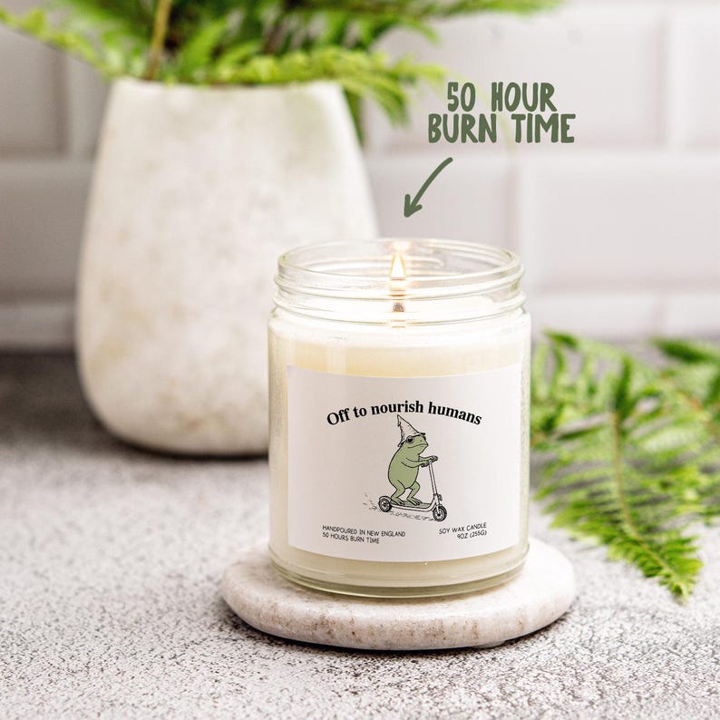 a candle labeled "Off to nourish humans" with a frog riding a scooter, placed on a white surface next to a plant.