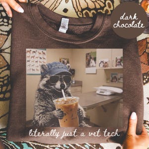 May include: A dark chocolate brown sweatshirt featuring a graphic print of a raccoon wearing a blue baseball cap and sunglasses while holding an iced coffee. The text below the image reads, "literally just a vet tech" in white script.