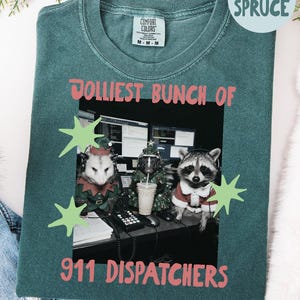 May include: A blue spruce coloured t-shirt featuring a graphic of an opossum, a skunk, and a raccoon wearing headsets and holiday attire at a desk. The text reads JOLLIEST BUNCH OF 911 DISPATCHERS, accented by light green star shapes on the fabric.