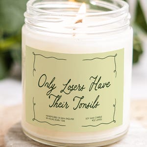 Könnte beinhalten: Eine brennende Sojawachskerze in einem durchsichtigen Glas. Die Kerze hat ein hellgrünes Etikett mit dem Text "Only Losers Have Their Tonsils". Das Etikett besagt auch "Handpoured in New England" und "50 Hours Burn Time". Die Kerze ist 255g.