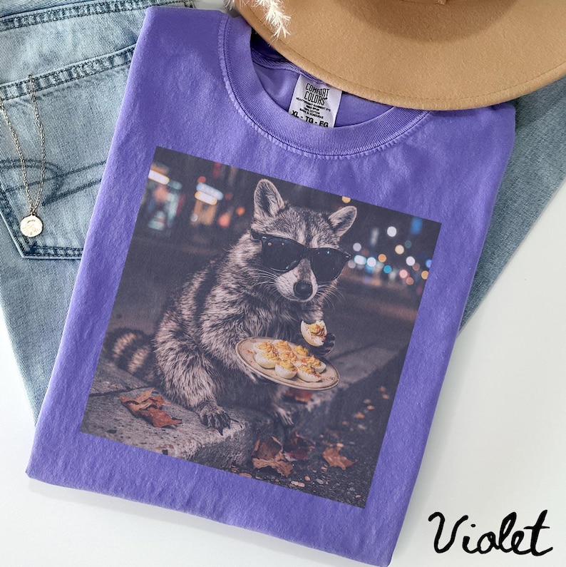 May include: A violet t-shirt featuring a graphic print of a raccoon wearing black sunglasses and eating deviled eggs from a plate. The shirt has a Comfort Colors label at the inner neckline, with the text XL, TG, EG visible on the tag.