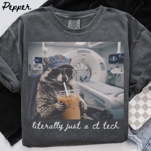 Op de afbeelding: Een antracietgrijs t-shirt met een afbeelding van een wasbeer die een baseballpet en zonnebril draagt en een ijskoffie vasthoudt. De achtergrond toont een CT-scanner in een medische ruimte. De tekst onderaan luidt, literally just a ct tech.