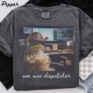 以下が含まれることがあります： チャコールグレーのTシャツ。カウボーイハットとサングラスを着用し、ヘッドセットを付けてアイスコーヒーを持っているカエルのグラフィックがプリントされています。グラフィックの下には白い筆記体で「wee woo dispatcher」という文字が書かれています。